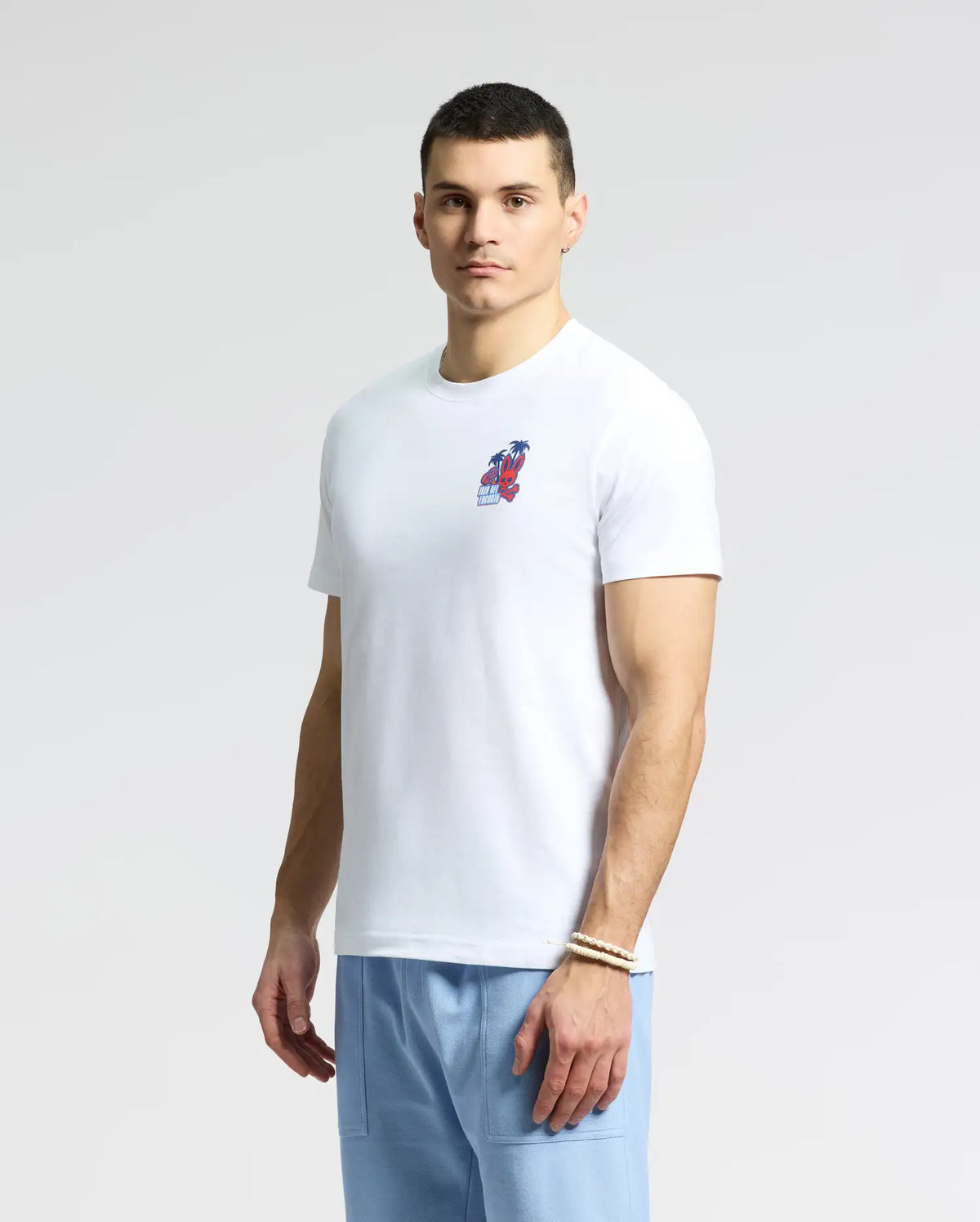 The individual dons a Psycho Bunny MENS PUERTO RICO TEE - B6U382F200 in white featuring a small, vibrant red and blue chest graphic, paired with light blue pants. Made from soft Pima cotton, it celebrates Puerto Rican pride. The background is plain and neutral as they face the camera directly.