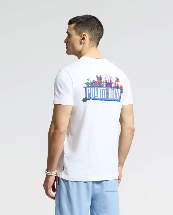 A man in a white Psycho Bunny Men's Puerto Rico Tee (B6U382F200) proudly displays his Puerto Rican pride with vibrant graphics on the back. He has short hair and wears light blue pants, standing out against a plain backdrop.