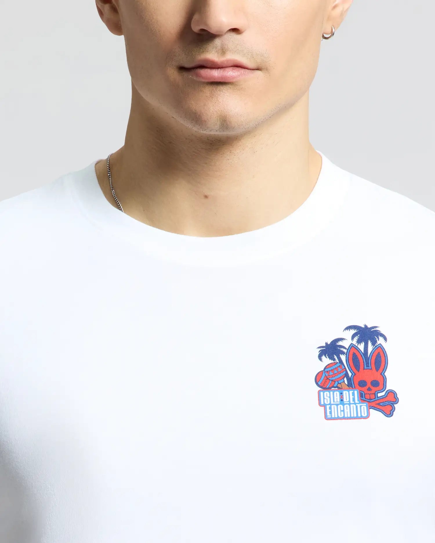 Wearing a Psycho Bunny Men's Puerto Rico Tee (B6U382F200) in white featuring palm trees, a long-eared creature, and "Isla del Encanto" in red and blue, paired with a thin silver necklace, exudes comfort and style—a subtle nod to Puerto Rican pride.