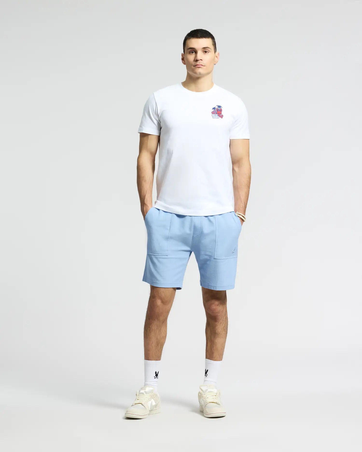 A person stands against a plain background wearing a Psycho Bunny MENS PUERTO RICO TEE - B6U382F200 with light blue shorts, white socks, and sneakers. They have their hands in pockets and maintain a neutral expression.