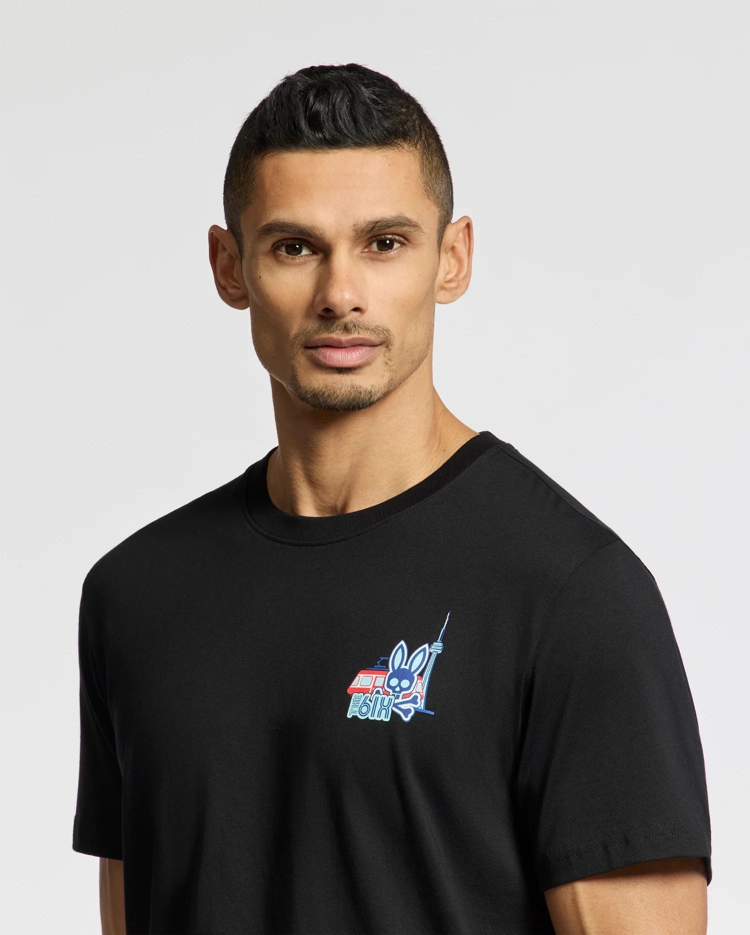 A man with short hair wears the Psycho Bunny MENS TORONTO TEE, a black shirt made from soft Pima cotton. It features a small, colorful bunny graphic and text on the upper left. The plain white background accentuates its striking yet subtle design.