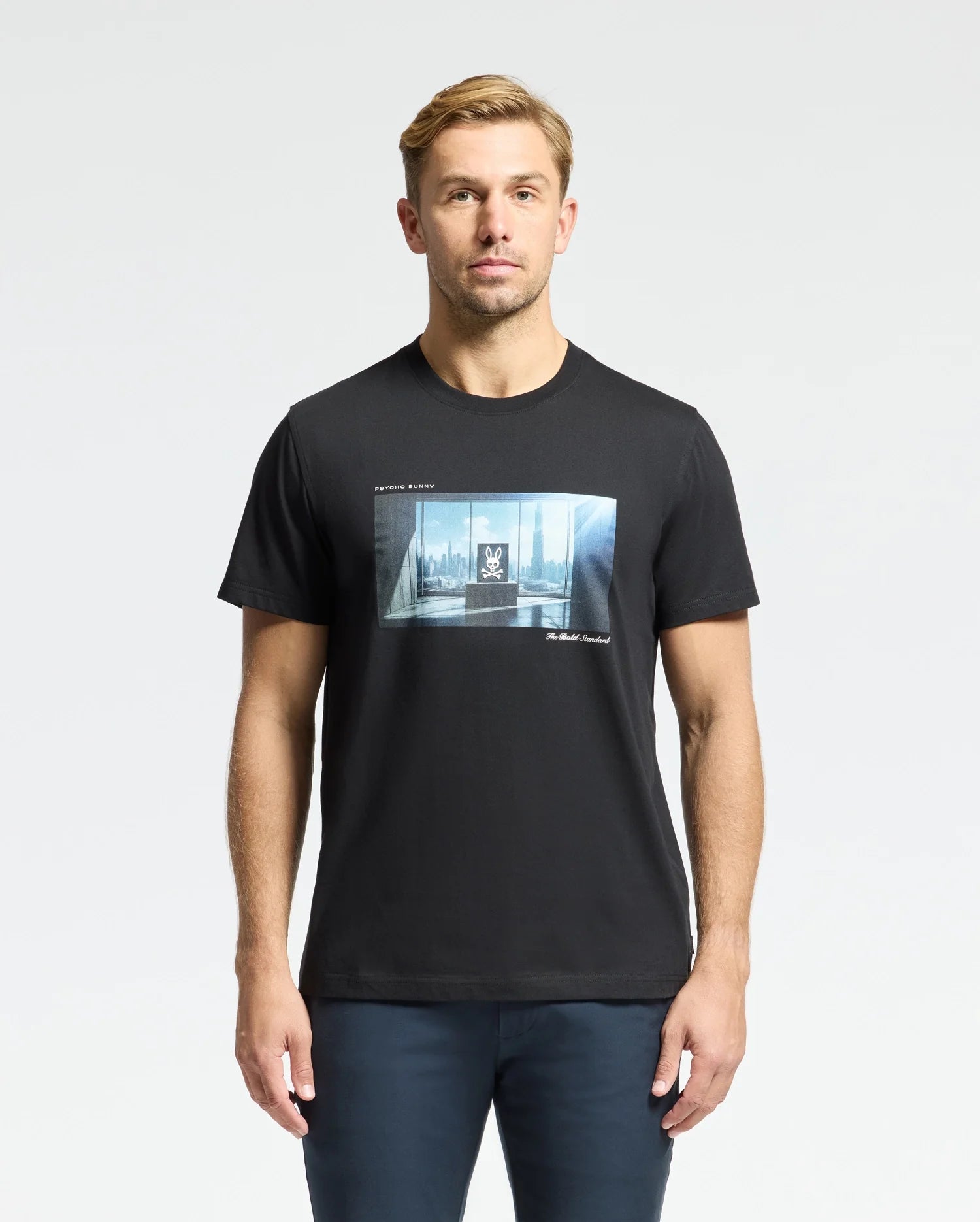 A man stands against a plain background wearing the Psycho Bunny MENS WARREN PHOTO REAL TEE - B6U388E200, a black Pima cotton T-shirt with a city skyline and window graphic. His short light brown hair complements his dark pants as he faces forward.