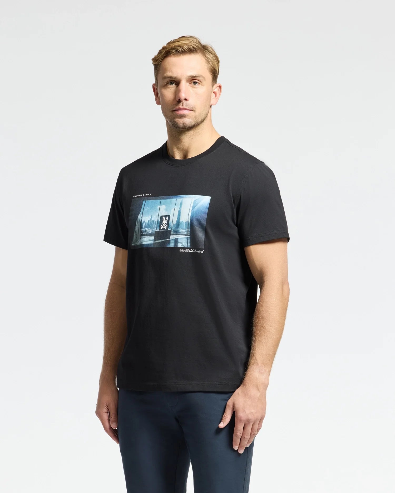 A man with light brown hair wears a Psycho Bunny Men's Warren Photo Real Tee, B6U388E200, made from Pima cotton with a city skyline graphic. The background is plain white.