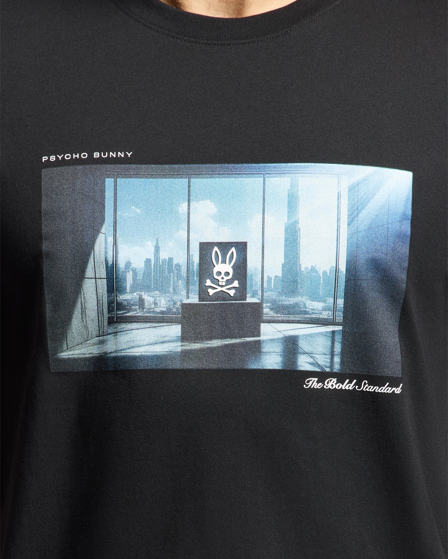 Wearing the Psycho Bunny MENS WARREN PHOTO REAL TEE, made from premium Pima cotton, a person stands in a room with large windows offering stunning city views. A screen highlights the bold Psycho Bunny skull logo and "The Bold Standard" text.