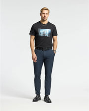 Against a plain background, a man wears a Psycho Bunny MENS WARREN PHOTO REAL TEE - B6U388E200 in black, navy pants, a black belt, and shoes. With short hair and a trimmed beard, he gazes into the camera with effortless style reminiscent of an urban city skyline.