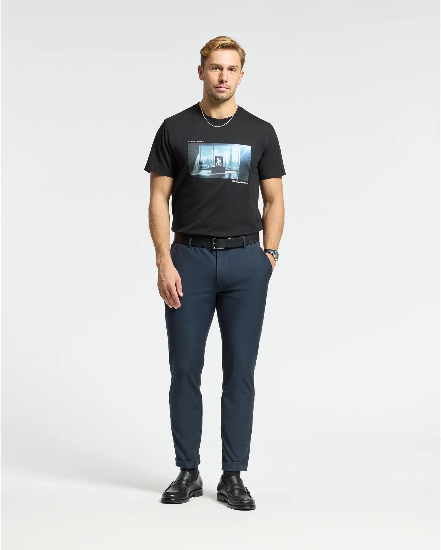 Against a plain background, a man wears a Psycho Bunny MENS WARREN PHOTO REAL TEE - B6U388E200 in black, navy pants, a black belt, and shoes. With short hair and a trimmed beard, he gazes into the camera with effortless style reminiscent of an urban city skyline.