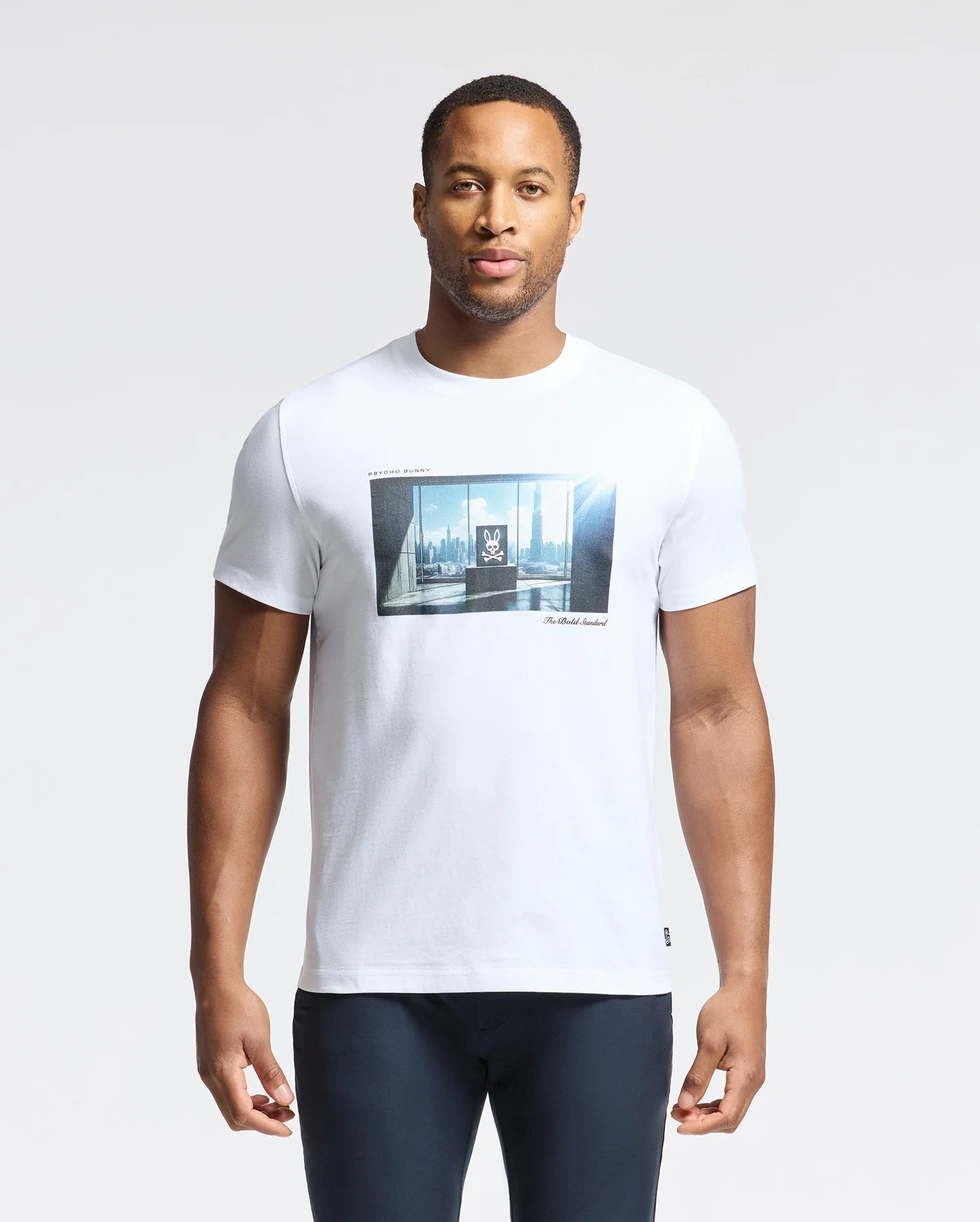 A person in a Psycho Bunny MENS WARREN PHOTO REAL TEE, made from soft Pima cotton jersey, showcases a city skyline window graphic while standing against a plain white background.