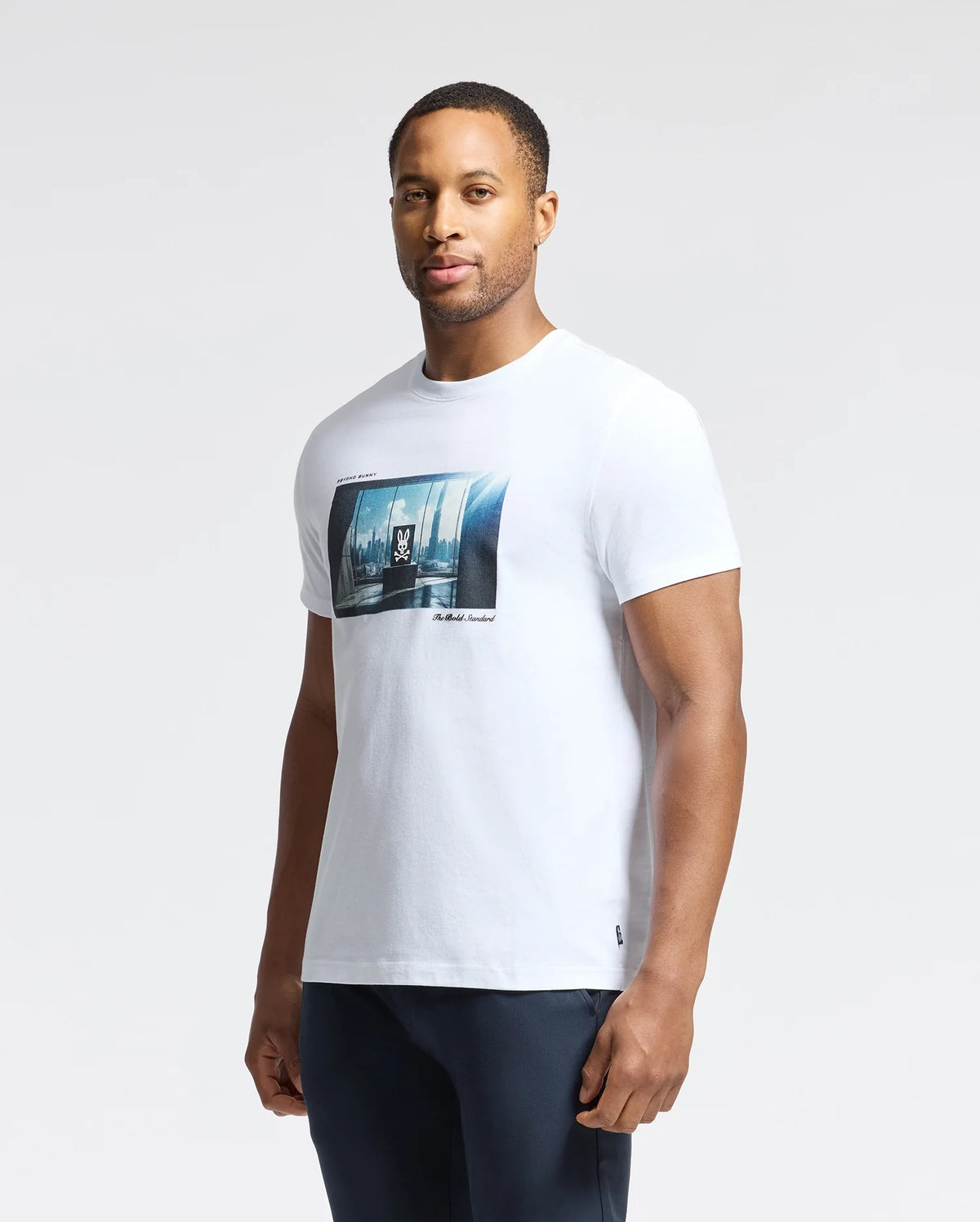 A person wears a MENS WARREN PHOTO REAL TEE - B6U388E200 by Psycho Bunny, made from Pima cotton jersey. The tee features a city skyline design with windows and a small logo. It's paired with dark pants against a white background.