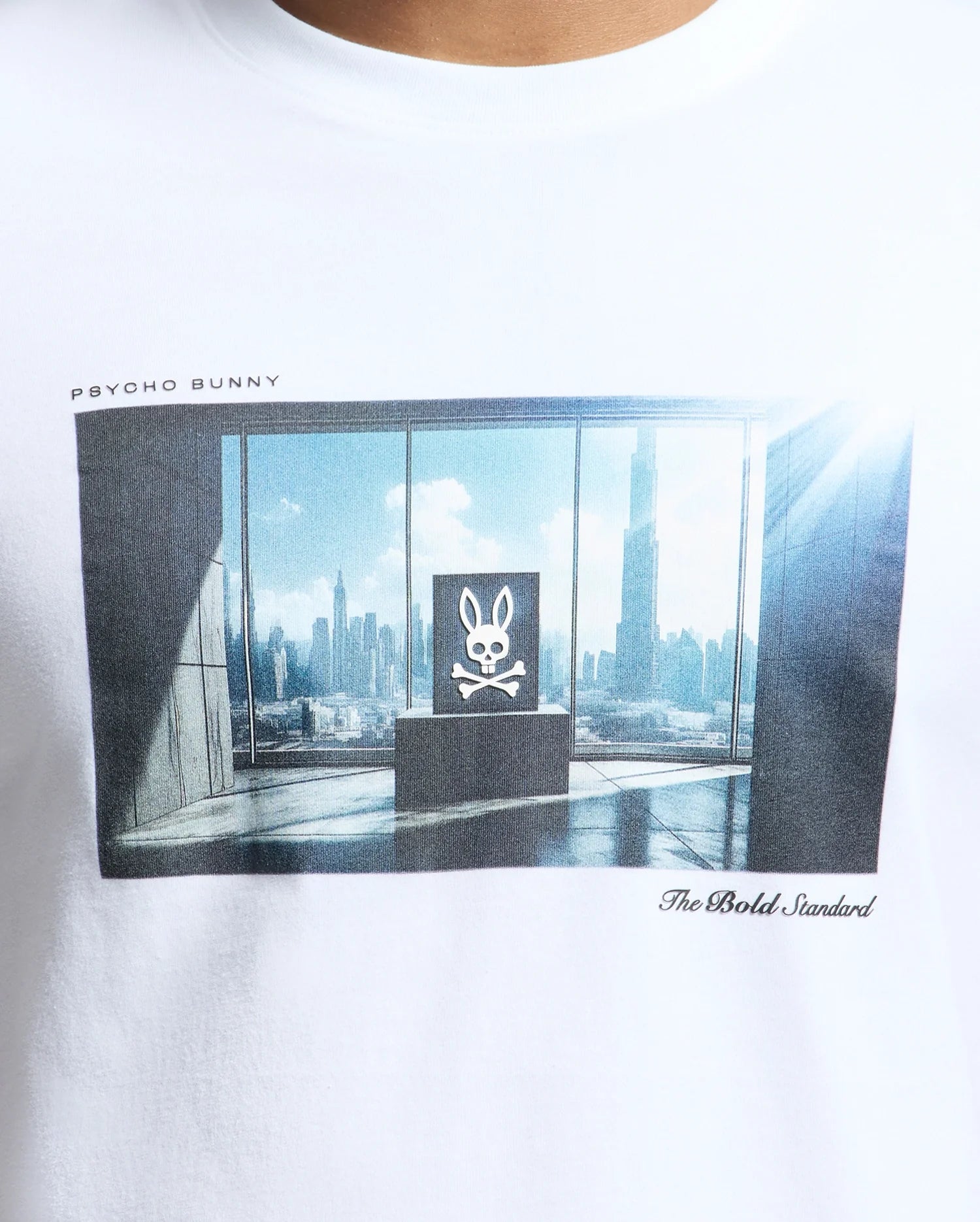 The MENS WARREN PHOTO REAL TEE - B6U388E200 by Psycho Bunny is a Pima cotton white T-shirt featuring a city skyline room print with a central skull and bunny ears. It displays "PSYCHO BUNNY" and "The Bold Standard.