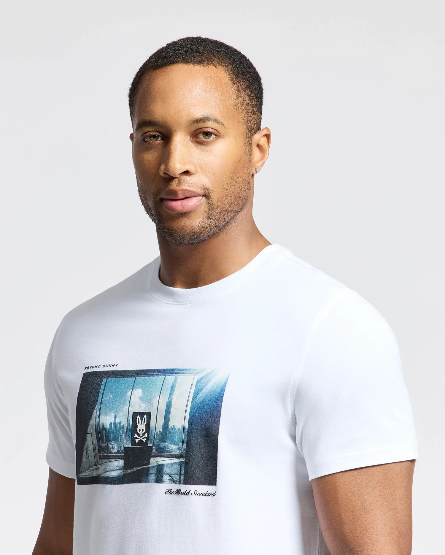 A man wears the Psycho Bunny MENS WARREN PHOTO REAL TEE (B6U388E200), crafted from soft Pima cotton jersey with a city skyline graphic through a window view, featuring a subtle rabbit logo on a plain light gray background.