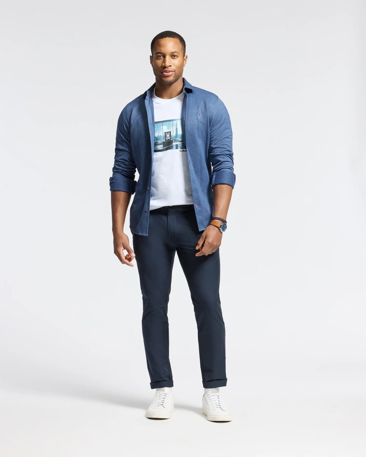 A man stands confidently on a plain white background, wearing a blue button-up shirt over a Psycho Bunny MENS WARREN PHOTO REAL TEE - B6U388E200 with a subtle city skyline design, paired with dark pants and white sneakers.