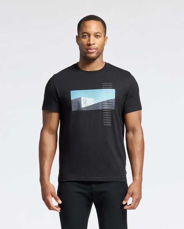 A man in the Psycho Bunny MENS NEIL PHOTO REAL TEE - B6U389E200, crafted from soft Pima cotton with a blue and white geometric gradient design, stands against a plain white background. His relaxed arms highlight the shirt's jersey fabric mirroring brutalist simplicity.