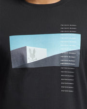 The Psycho Bunny Men's Neil Photo Real Tee (B6U389E200) features a black Pima cotton fabric with a rectangular graphic displaying a cube and bunny skull silhouette against a light blue sky. The brutalist-style design repeats vertically alongside the text "Psycho Bunny.