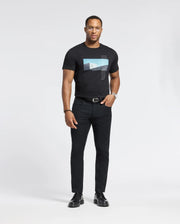 A man with short hair and glasses, wearing a Psycho Bunny MENS NEIL PHOTO REAL TEE - B6U389E200 made of soft Pima cotton, is paired with black jeans and shoes. He has a watch on his left wrist and looks confidently at the camera against a plain background, hands by his sides.
