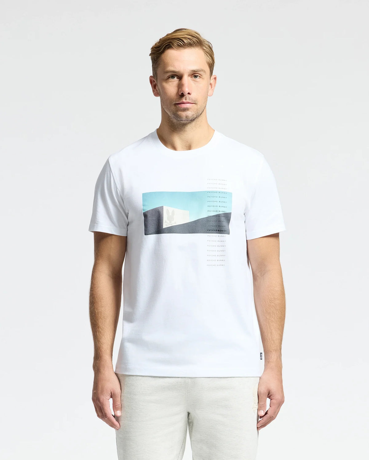 A man with short blond hair wears a Psycho Bunny MENS NEIL PHOTO REAL TEE - B6U389E200, crafted from luxurious Peruvian Pima cotton, featuring blue and black abstract shapes. He stands against a plain white background.