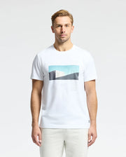 A man with short blond hair wears a Psycho Bunny MENS NEIL PHOTO REAL TEE - B6U389E200, crafted from luxurious Peruvian Pima cotton, featuring blue and black abstract shapes. He stands against a plain white background.