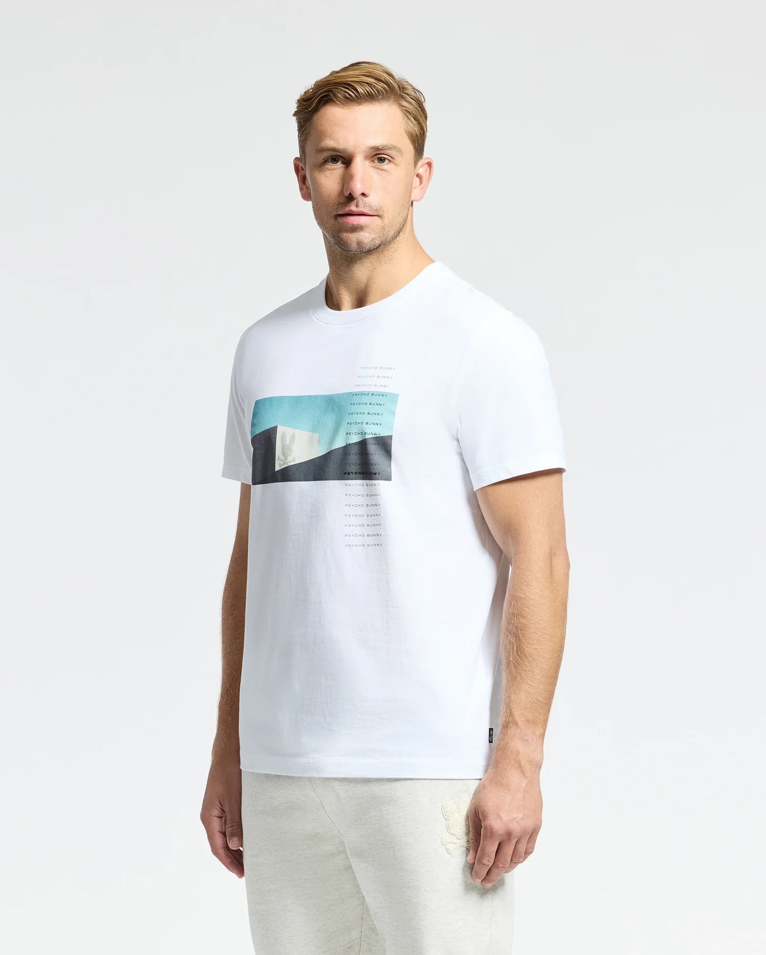 A man with short blond hair wears a Psycho Bunny MENS NEIL PHOTO REAL TEE, crafted from soft Peruvian Pima cotton with a rectangular graphic print on the front against a plain background.