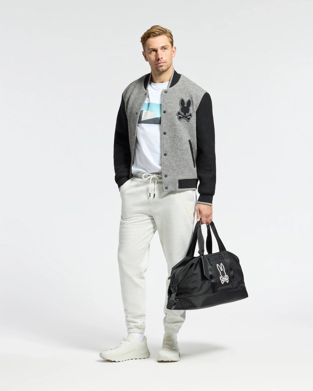 A person stands against a plain background wearing a gray and black varsity jacket, Psycho Bunny MENS NEIL PHOTO REAL TEE in premium Peruvian Pima cotton, light gray joggers, and white sneakers. They're holding a black duffel bag with a white logo in one hand.
