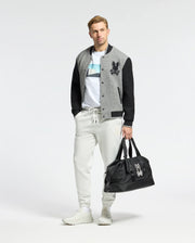 A person stands against a plain background wearing a gray and black varsity jacket, Psycho Bunny MENS NEIL PHOTO REAL TEE in premium Peruvian Pima cotton, light gray joggers, and white sneakers. They're holding a black duffel bag with a white logo in one hand.