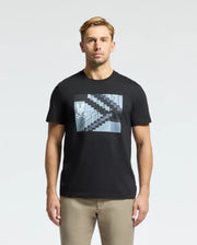 A man with short light brown hair wears a Psycho Bunny MENS DEAN PHOTO REAL TEE - B6U390E200 in black Pima cotton jersey, featuring a rabbit graphic amid urban geometric shapes on a staircase, facing forward against a plain white background.