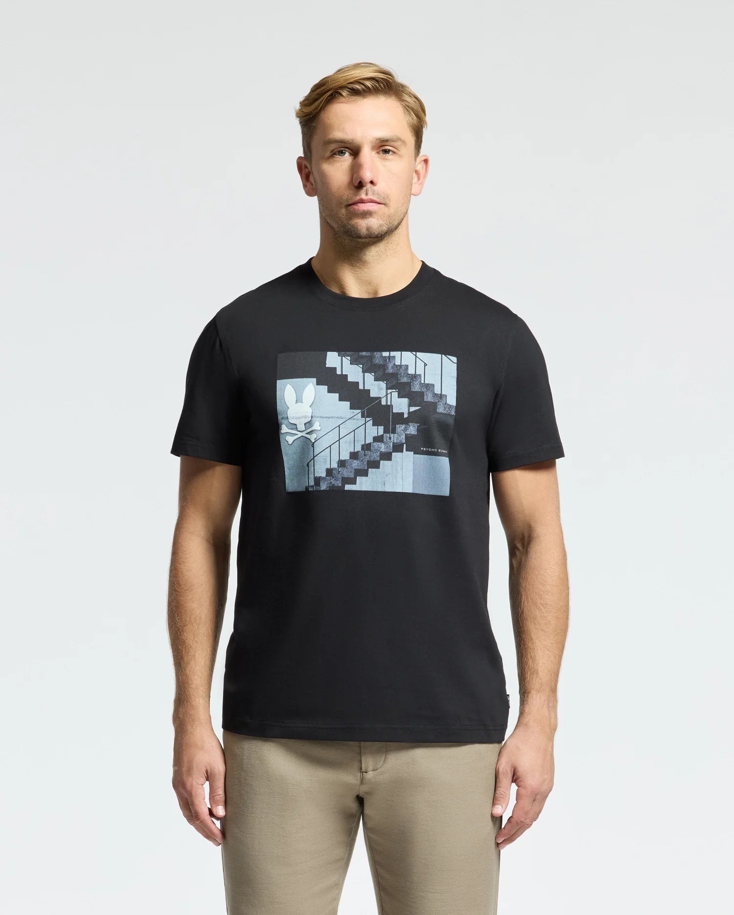 A man with short light brown hair wears a Psycho Bunny MENS DEAN PHOTO REAL TEE - B6U390E200 in black Pima cotton jersey, featuring a rabbit graphic amid urban geometric shapes on a staircase, facing forward against a plain white background.
