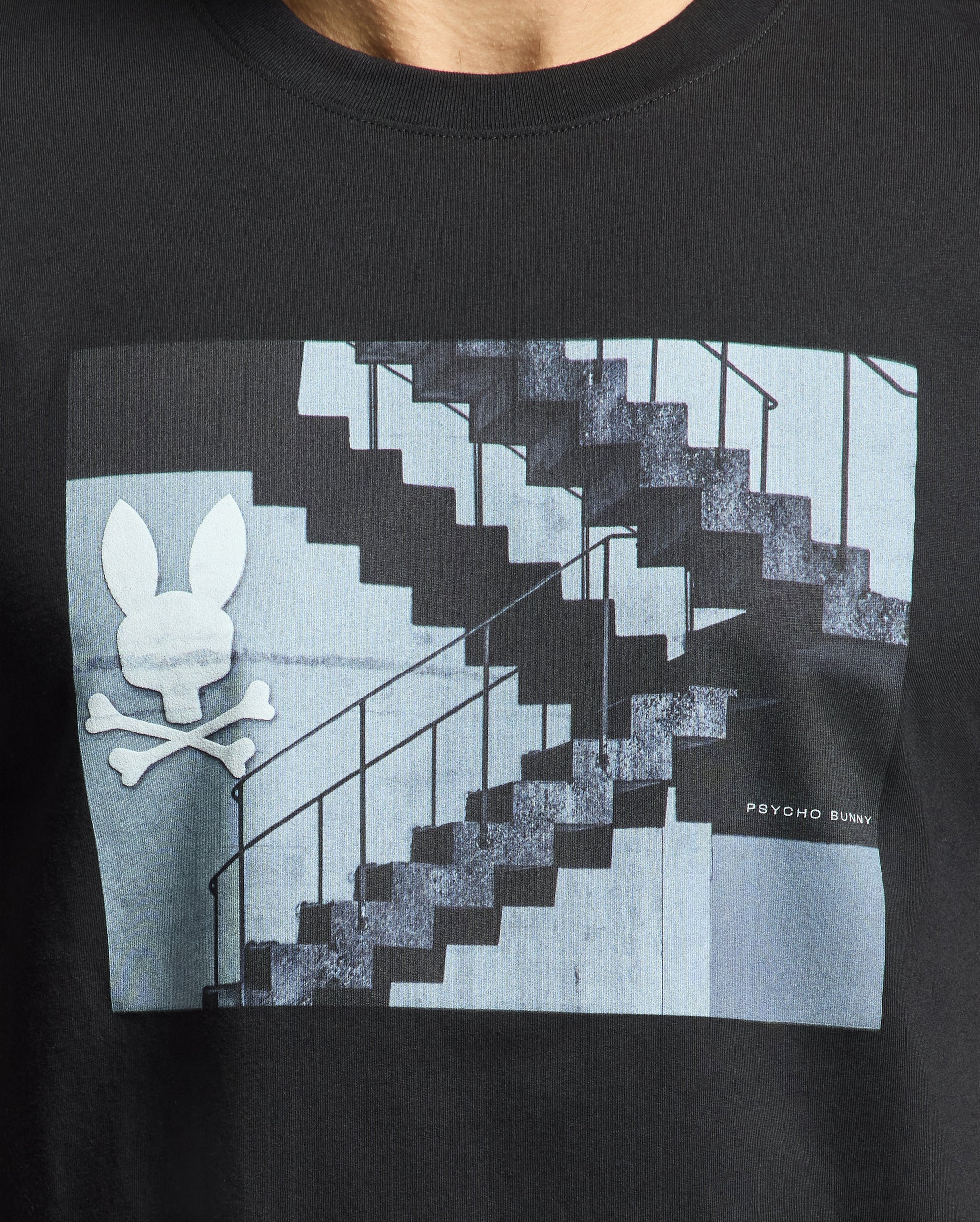 The Men's Dean Photo Real Tee by Psycho Bunny is a stylish Pima cotton jersey featuring an urban geometric shapes graphic with a dual staircase pattern, a bunny-headed skull and crossbones on the left, and "Psycho Bunny" text on the bottom right.