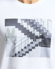 A person wears the Mens Dean Photo Real Tee by Psycho Bunny, made from soft Pima cotton. It boasts a white design with urban geometric shapes and features the bunny skull and crossbones logo on the lower right corner alongside the brand name.