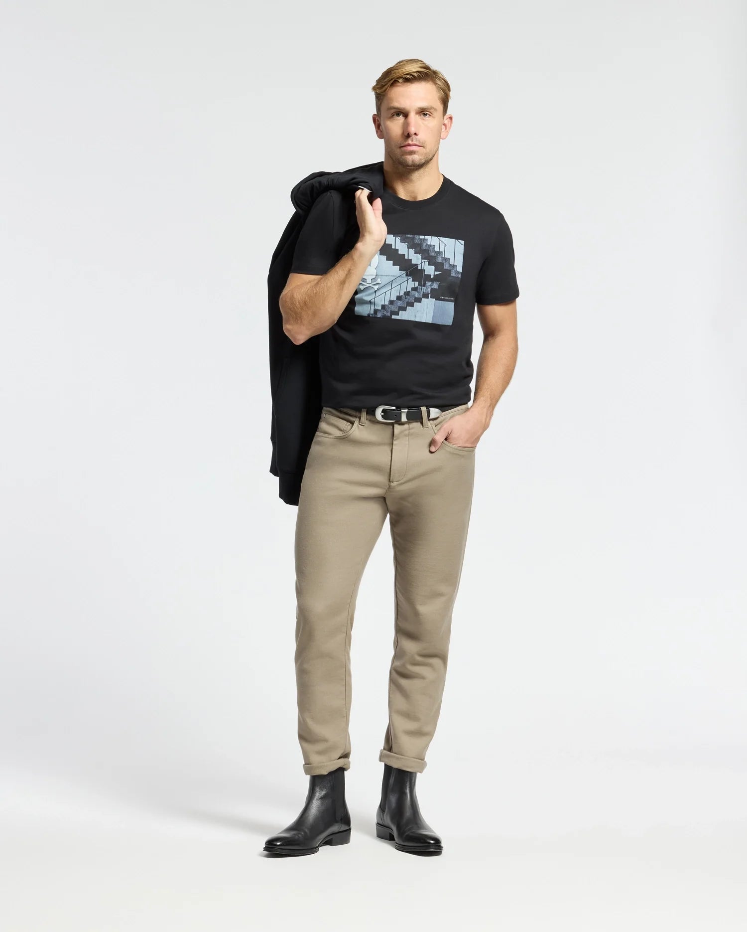 A man confidently wears a Psycho Bunny MENS DEAN PHOTO REAL TEE (B6U390E200), showcasing its urban geometric design. Styled with beige pants and black boots, he casually drapes a jacket over his shoulder, hand in pocket, posing coolly against a plain white background.