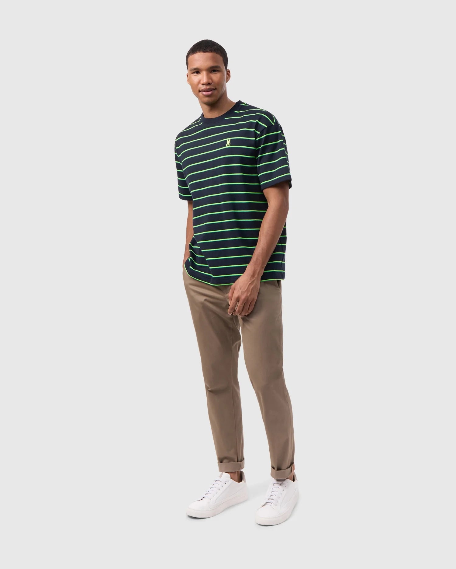 A person stands against a plain white background, smiling. They wear the Psycho Bunny MENS ALTON STRIPE OVERSIZED TEE - B6U409C200 with 90s-inspired two-tone stripes in dark green and black, beige pants, and white sneakers. Their hands are casually tucked into their pockets.