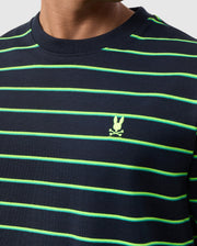 Close-up of a person wearing the Psycho Bunny MENS ALTON STRIPE OVERSIZED TEE - B6U409C200 in a navy blue and green striped shirt in a relaxed fit, featuring 90s-inspired two-tone stripes and a small embroidered yellow skull and crossbones logo on the chest. The person's face is not visible.