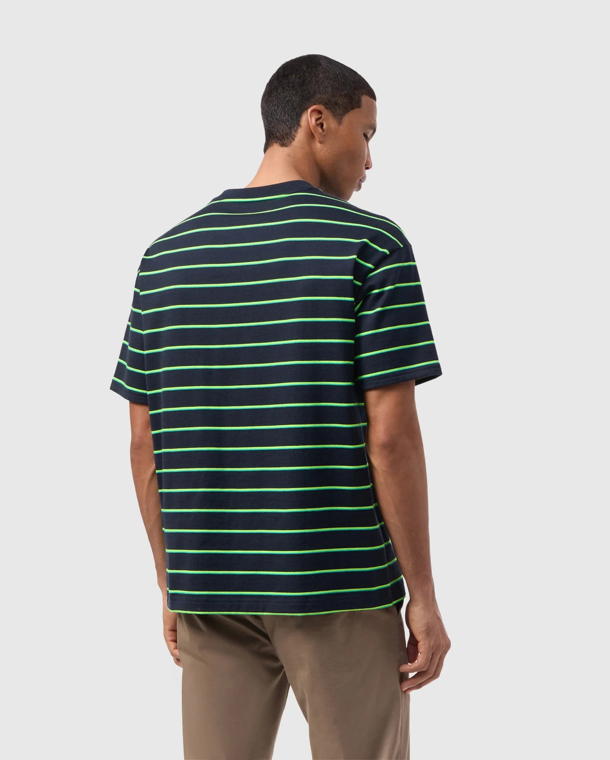 A person with short hair is standing and facing away from the camera, wearing a Psycho Bunny MENS ALTON STRIPE OVERSIZED TEE - B6U409C200 with 90s-inspired green two-tone stripes and khaki pants. The relaxed fit and heavyweight jersey fabric add a touch of timeless style. The background is plain white or light grey.