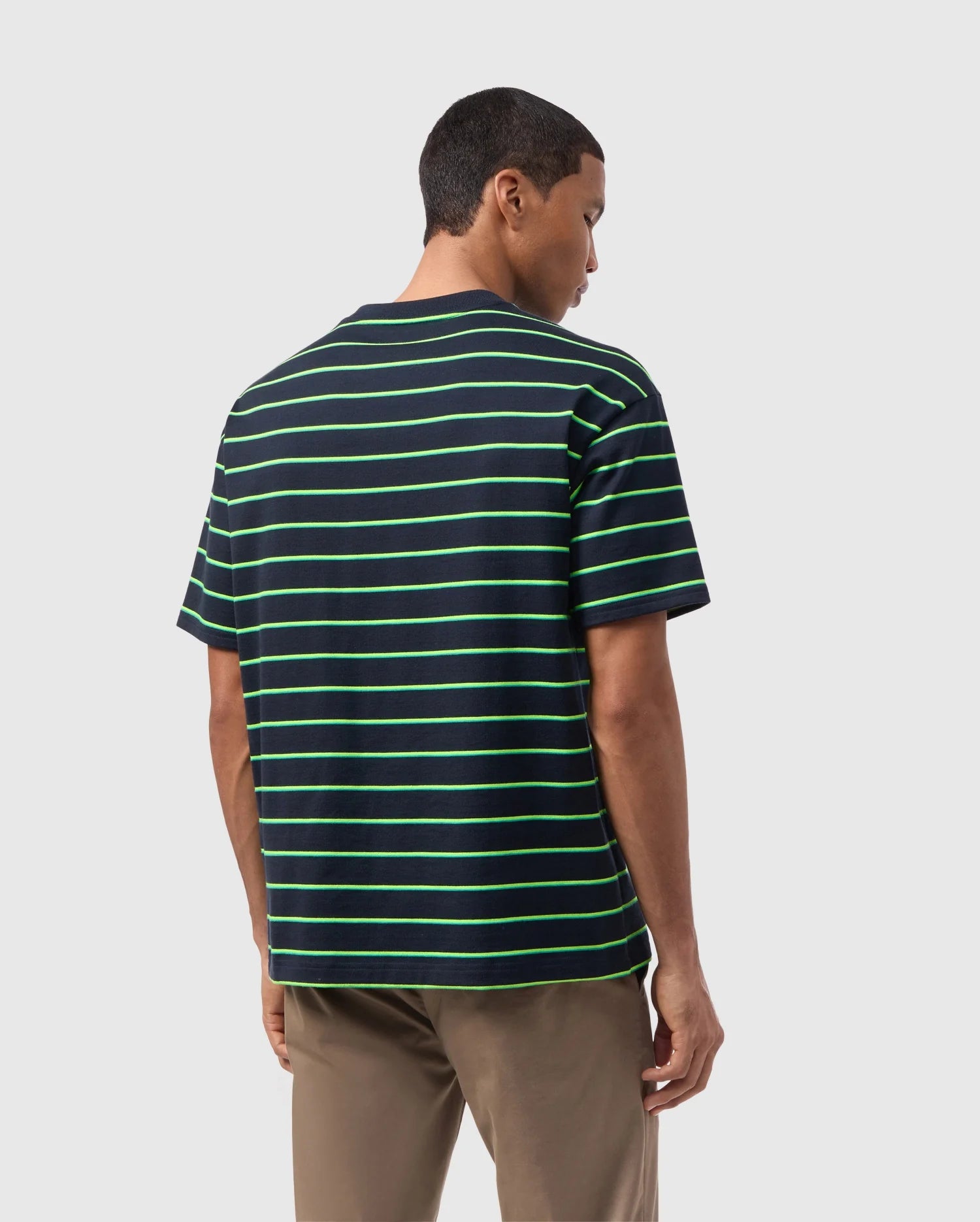 A person with short hair is standing and facing away from the camera, wearing a Psycho Bunny MENS ALTON STRIPE OVERSIZED TEE - B6U409C200 with 90s-inspired green two-tone stripes and khaki pants. The relaxed fit and heavyweight jersey fabric add a touch of timeless style. The background is plain white or light grey.