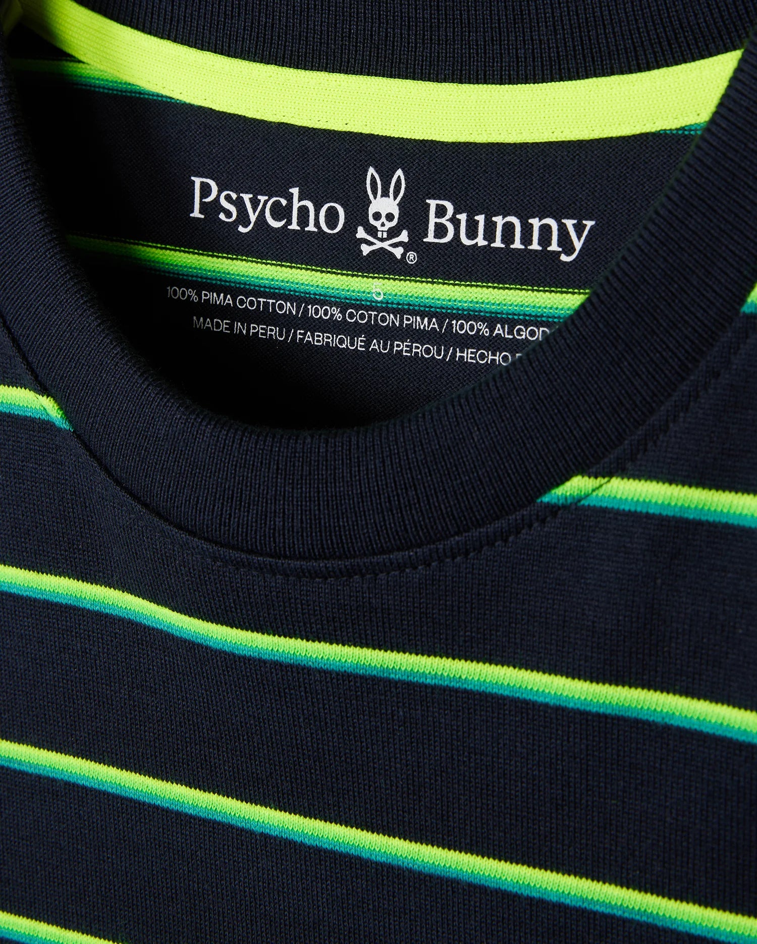 Close-up of a navy blue MENS ALTON STRIPE OVERSIZED TEE - B6U409C200 featuring 90s-inspired two-tone neon yellow and green horizontal stripes. The relaxed fit shirt's label inside the collar shows "Psycho Bunny" with a bunny skull logo, indicating it’s made of 100% Pima cotton and crafted in Peru.
