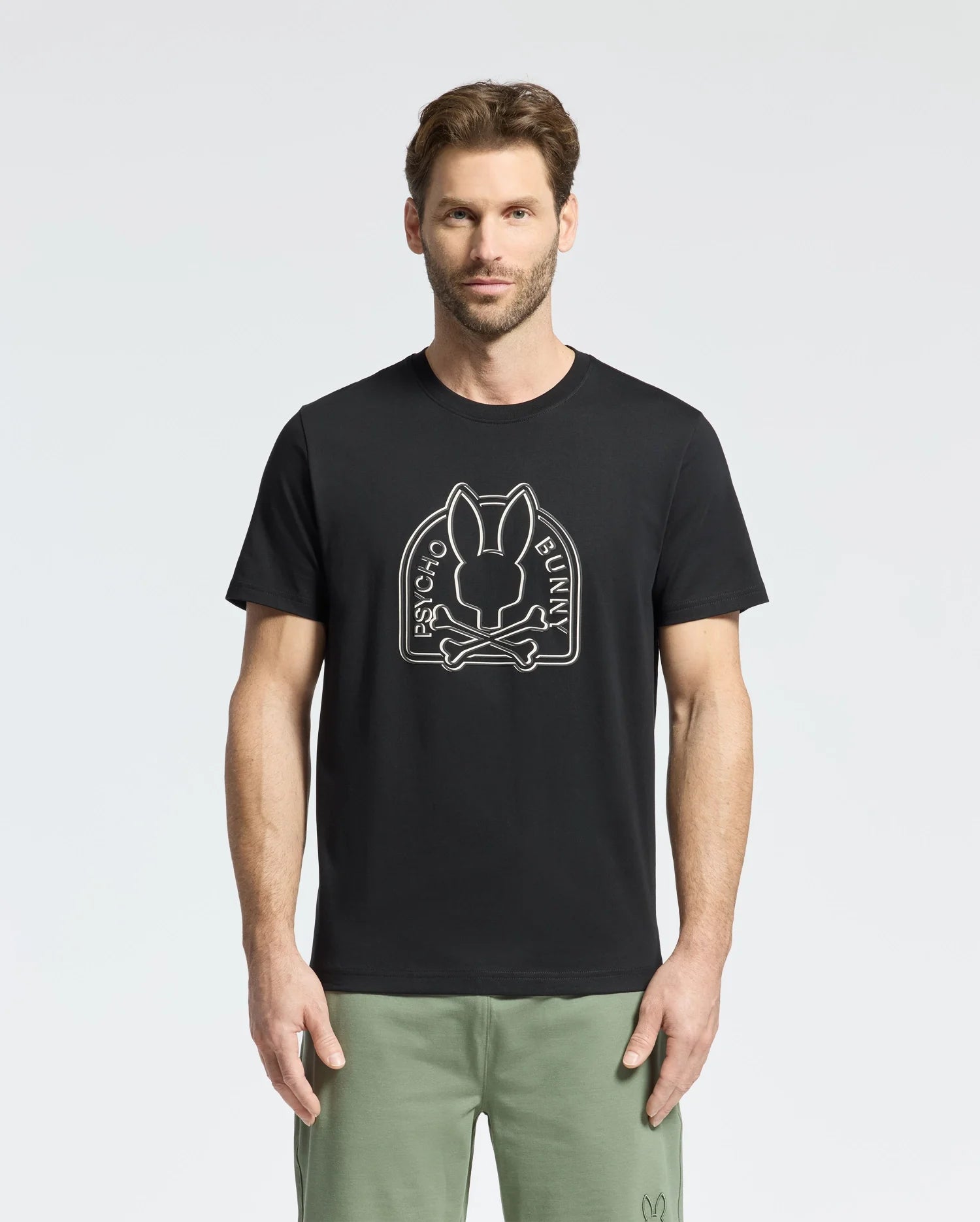 A man wears a Psycho Bunny MENS MELBOURNE GRAPHIC TEE in black, showcasing a Bunny graphic and text. He pairs it with light green pants against a plain white backdrop.
