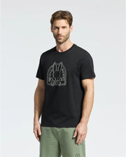 A man wears a Psycho Bunny MENS MELBOURNE GRAPHIC TEE - B6U410F200, featuring a bunny with crossbones graphic and "Psycho Bunny" text. He pairs it with light green pants against a plain, neutral background.
