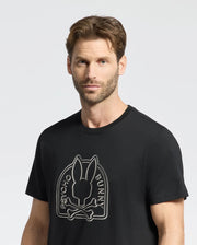 A short brown-haired, bearded man stands against a plain light background, wearing the Psycho Bunny MENS MELBOURNE GRAPHIC TEE - B6U410F200 featuring Peruvian Pima knit with a bold Bunny graphic and crossbones design.