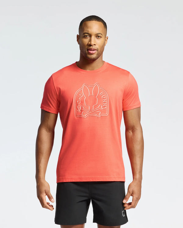 A man stands against a white background wearing Psycho Bunny's MENS MELBOURNE GRAPHIC TEE in red, crafted from soft, eco-friendly Peruvian Pima Cotton, featuring a charming bunny design and paired with black shorts.