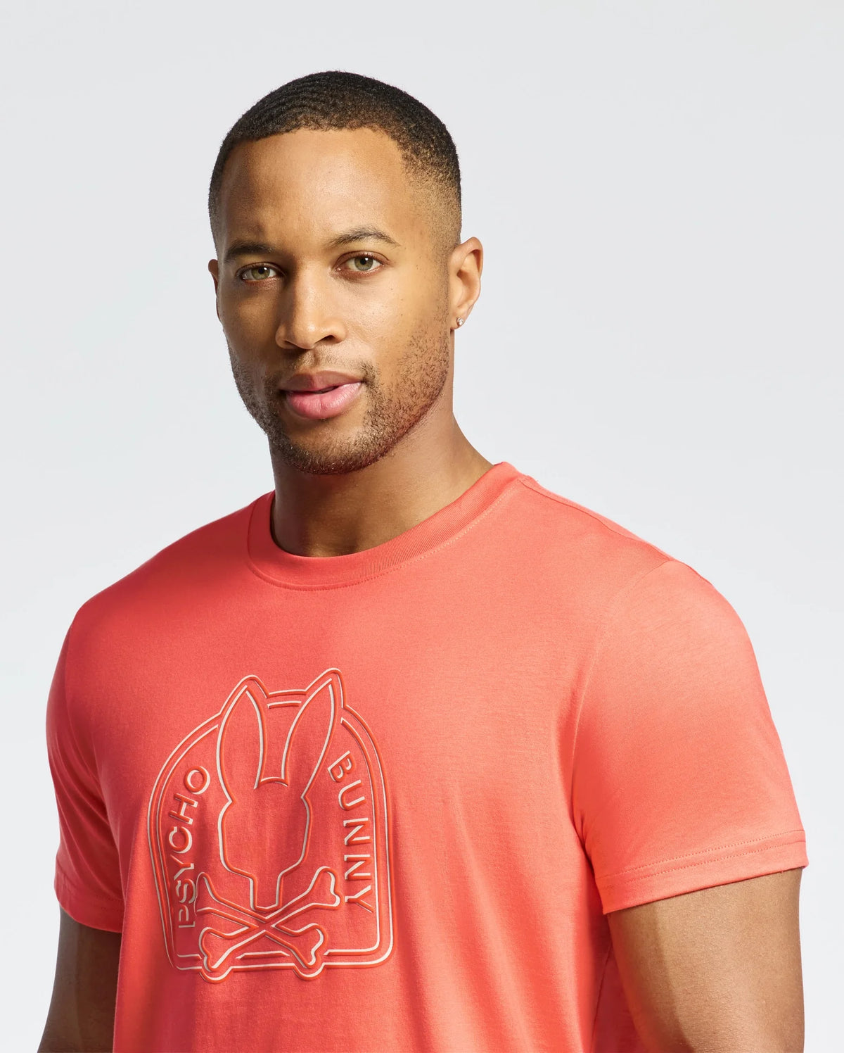 A person in a coral Psycho Bunny's MENS MELBOURNE GRAPHIC TEE - B6U410F200 made from eco-friendly Peruvian Pima Cotton stands against a plain background. The T-shirt showcases a stylized bunny graphic logo and text, perfectly complementing their short hair.