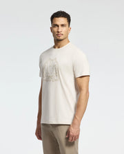 A man wears a beige Psycho Bunny MENS MELBOURNE GRAPHIC TEE crafted from luxurious Peruvian Pima cotton, featuring a large stylized rabbit embroidery. Paired with matching beige pants, the eco-friendly style stands out against the white background, highlighting his casual attire.