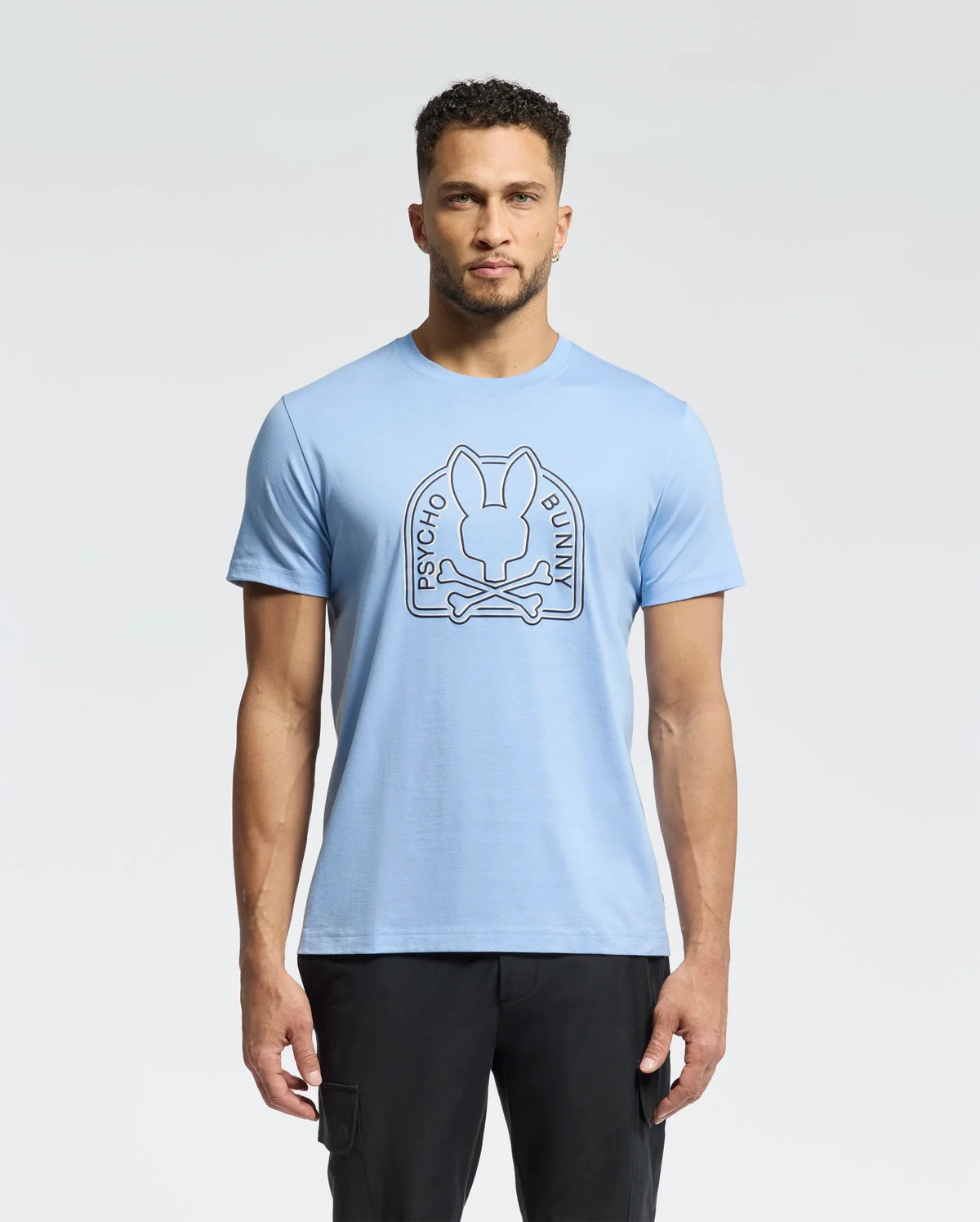 A person with short curly hair wears a Psycho Bunny MENS MELBOURNE GRAPHIC TEE - B6U410F200, a light blue tee featuring a bunny and crossbones graphic. They stand against a white backdrop in black pants. The tee’s soft Peruvian Pima cotton offers comfort to its edgy design.