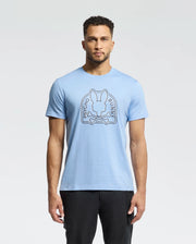 A person with short curly hair wears a Psycho Bunny MENS MELBOURNE GRAPHIC TEE - B6U410F200, a light blue tee featuring a bunny and crossbones graphic. They stand against a white backdrop in black pants. The tee’s soft Peruvian Pima cotton offers comfort to its edgy design.