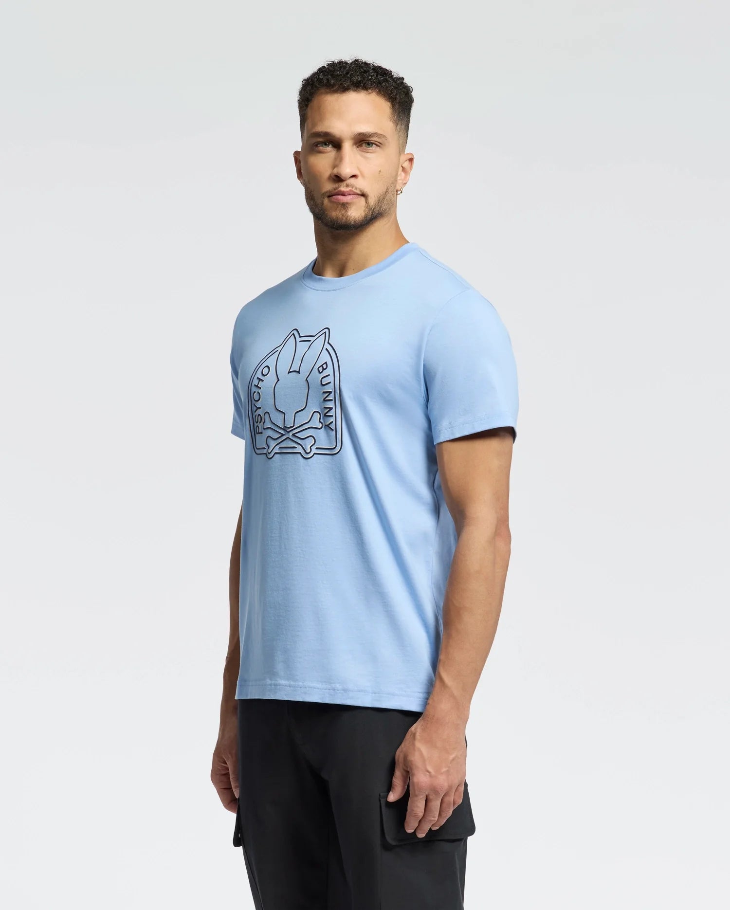A man in a light blue Psycho Bunny MENS MELBOURNE GRAPHIC TEE (B6U410F200), with a bunny graphic and text, is paired with black pants against a plain light background.
