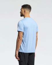 A man with short curly hair wears a light blue MENS MELBOURNE GRAPHIC TEE by Psycho Bunny, featuring a subtle bunny graphic, paired with black pants. He stands facing away against a plain, light background.