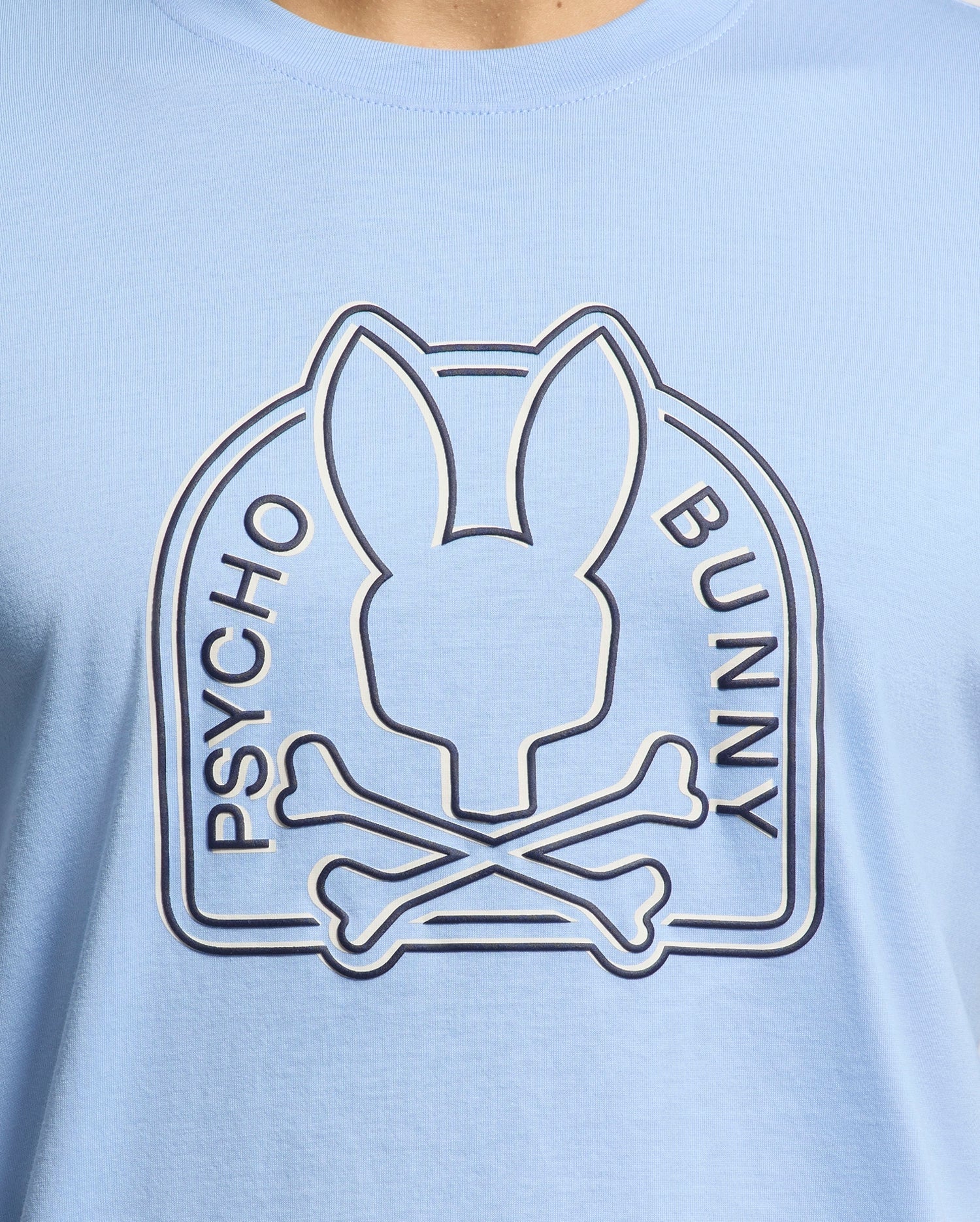 The MENS MELBOURNE GRAPHIC TEE from Psycho Bunny, style B6U410F200, features a light blue design with a bunny head silhouette above crossed bones, encircled by "PSYCHO BUNNY," crafted from luxurious Peruvian Pima cotton.