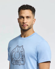 A man with short curly hair poses against a plain background wearing the light blue Psycho Bunny Mens Melbourne Graphic Tee, made from luxurious Peruvian Pima cotton and featuring a stylized bunny graphic.
