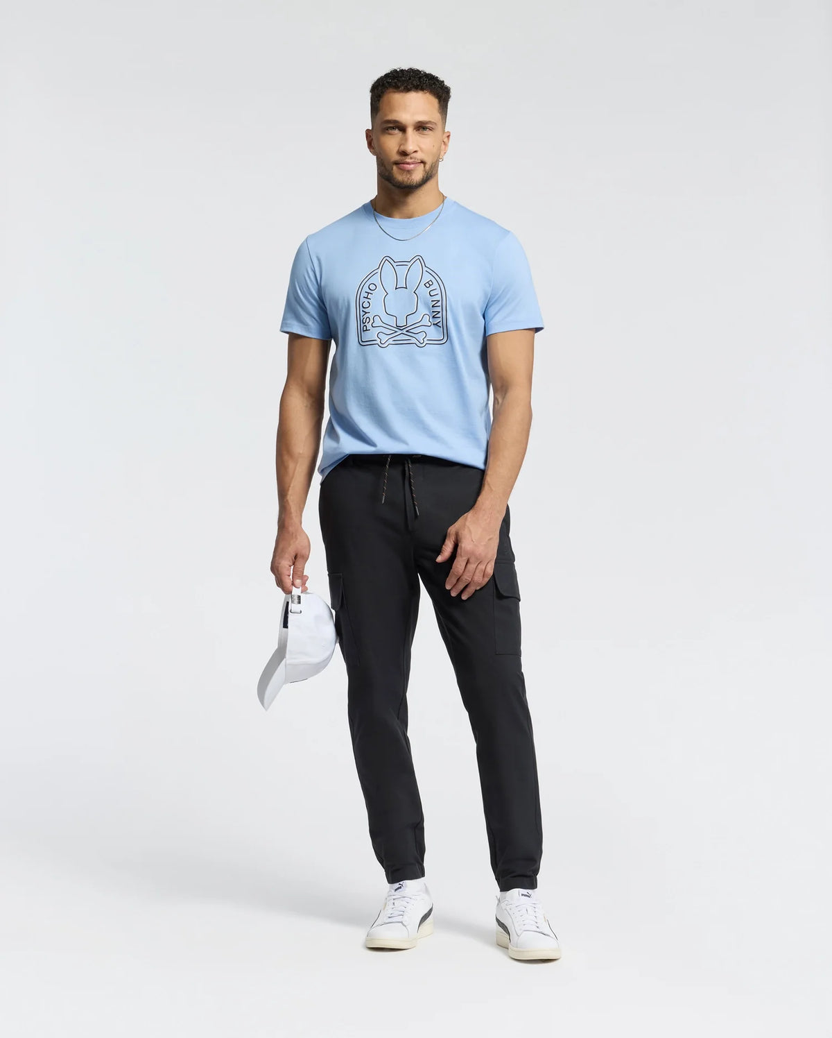 A man in a light blue Psycho Bunny Melbourne tee, B6U410F200, with a bunny graphic, black pants, and white sneakers holds a white cap. He stands against a plain white background, effortlessly showcasing the casual charm of Peruvian Pima cotton.