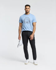 A man in a light blue Psycho Bunny Melbourne tee, B6U410F200, with a bunny graphic, black pants, and white sneakers holds a white cap. He stands against a plain white background, effortlessly showcasing the casual charm of Peruvian Pima cotton.