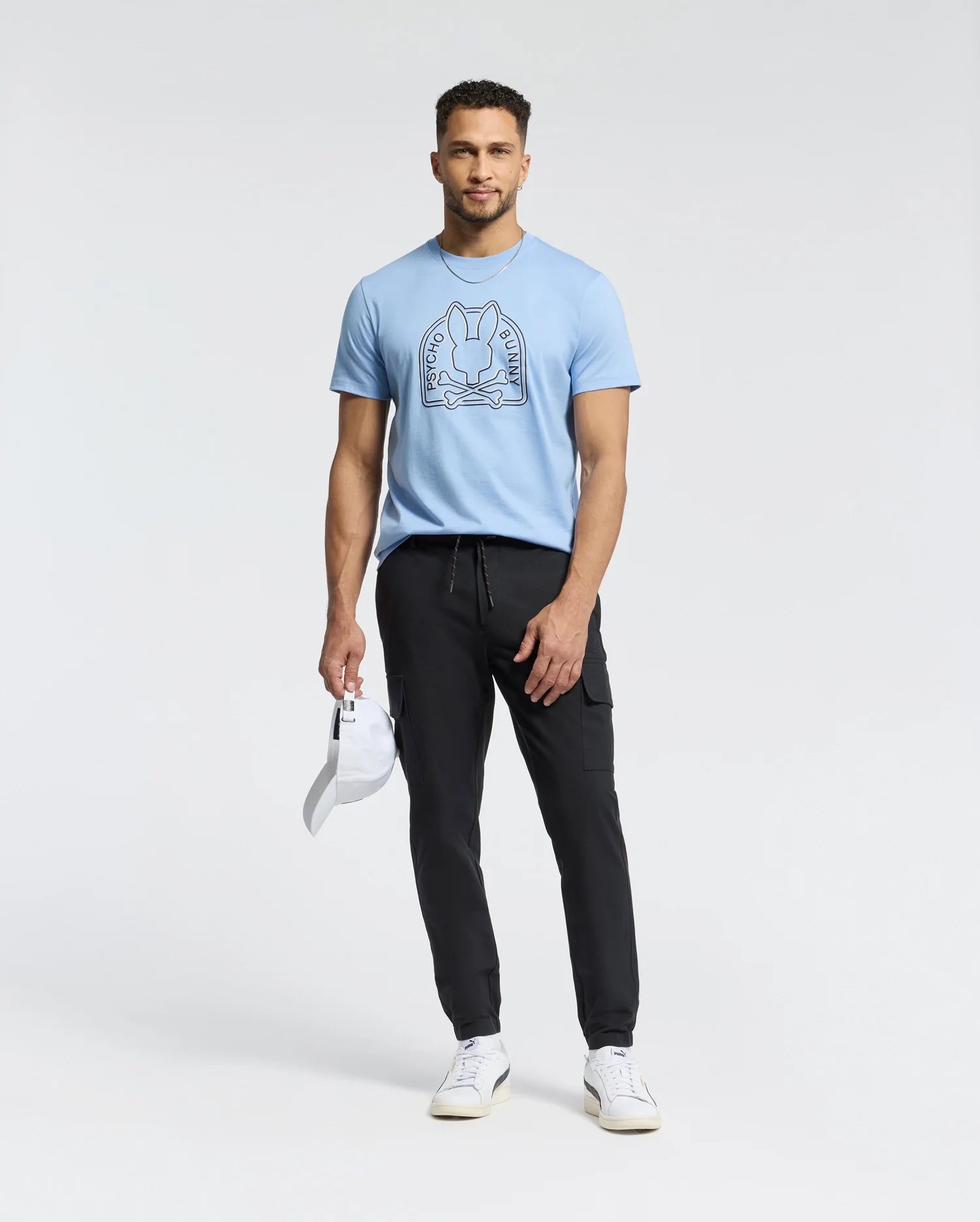 A man in a light blue Psycho Bunny Melbourne tee, B6U410F200, with a bunny graphic, black pants, and white sneakers holds a white cap. He stands against a plain white background, effortlessly showcasing the casual charm of Peruvian Pima cotton.