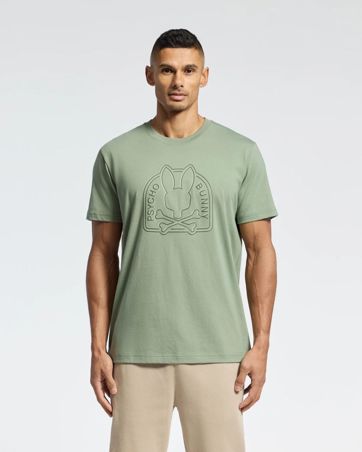 A man stands against a white backdrop wearing the eco-friendly Psycho Bunny Men's Melbourne Graphic Tee in light green, featuring a bunny and crossbones logo. It's crafted from luxurious Peruvian Pima cotton and paired with beige pants for a stylish look.