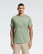 A man stands against a white backdrop wearing the eco-friendly Psycho Bunny Men's Melbourne Graphic Tee in light green, featuring a bunny and crossbones logo. It's crafted from luxurious Peruvian Pima cotton and paired with beige pants for a stylish look.
